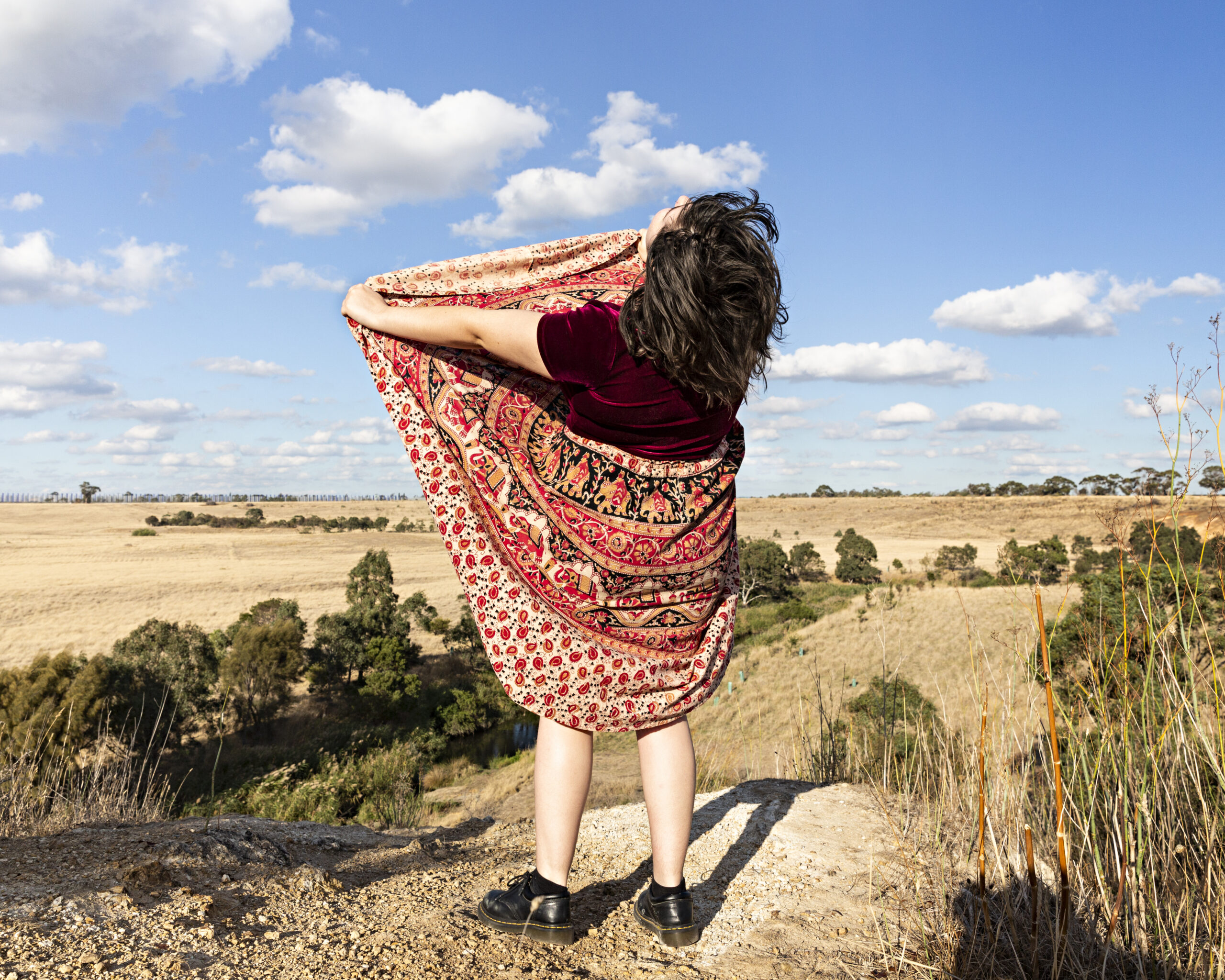 A photo of a person wearing a dress standing above a rock. The person has their back to the camera and is lifting the front of their dress up.