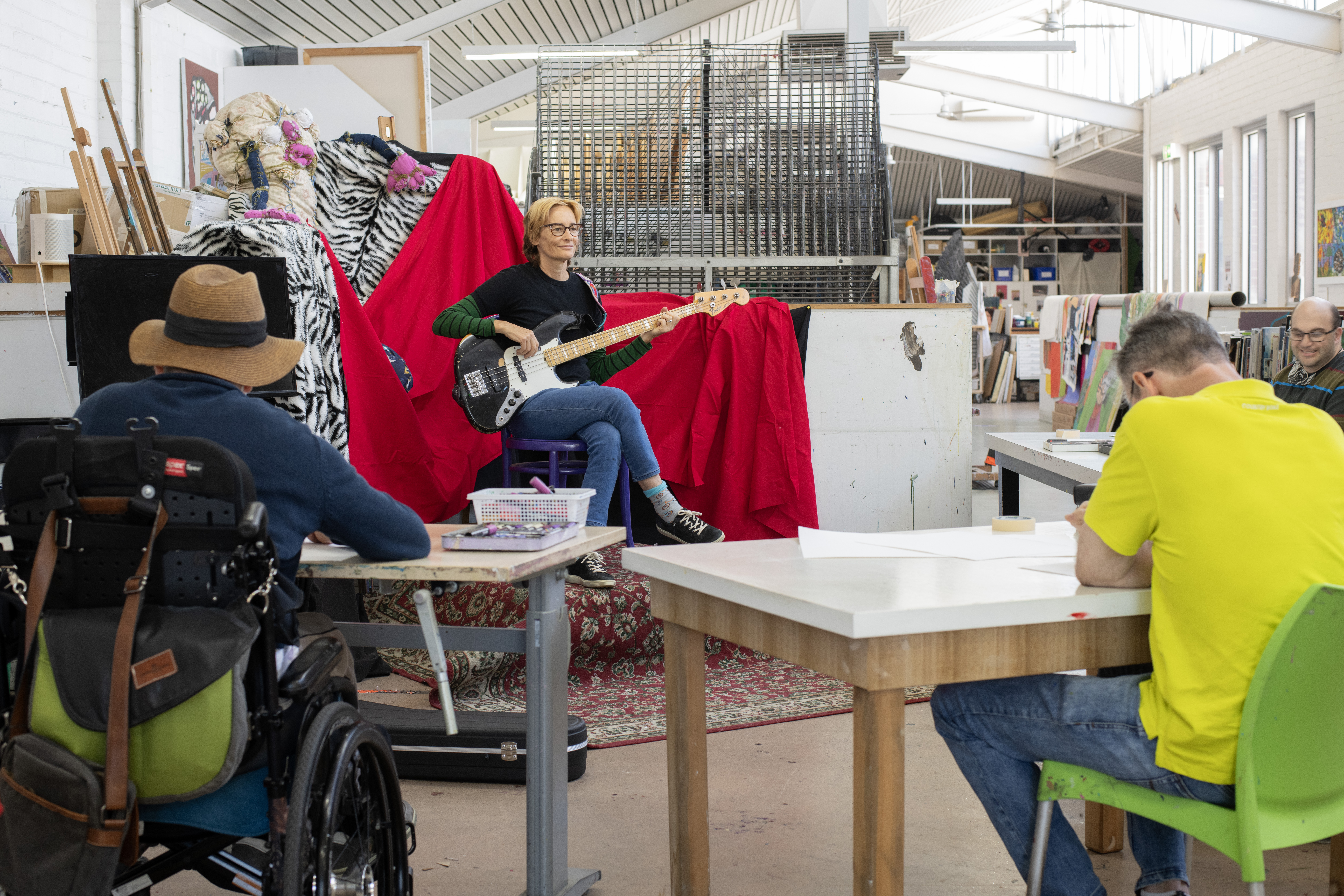 Janet English sits with her bass guitar on her lap in the Arts Project Australia studio while artists draw her portrait.